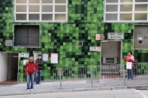The green wall drew me in first, its pixel-like pattern feeling oddly alive against the street. As people settled into place, their distance and mannequin-like gestures made the scene feel almost staged. The building’s function adds another layer, grounding the image while quietly complicating it.