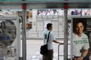 Captured in Tsim Sha Tsui, one of the most heavily advertised areas in Hong Kong, this scene unfolded at a bus stop framed by competing images. A middle aged man stood quietly, appearing unaware of the fashion billboard across the street. Positioned between two larger than life faces, he seemed momentarily caught in their gaze. The advertisements pressed in from both sides, while he remained still, holding his place in a space where scale, attention, and presence rarely belong to the same figure.